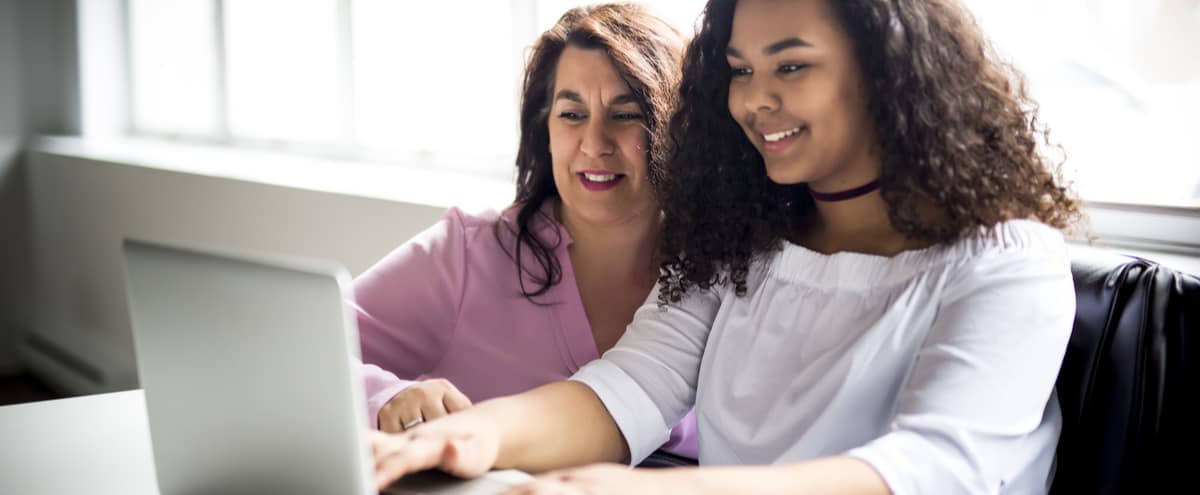 A happy mother and daughter sit at a desk working on a laptop together.